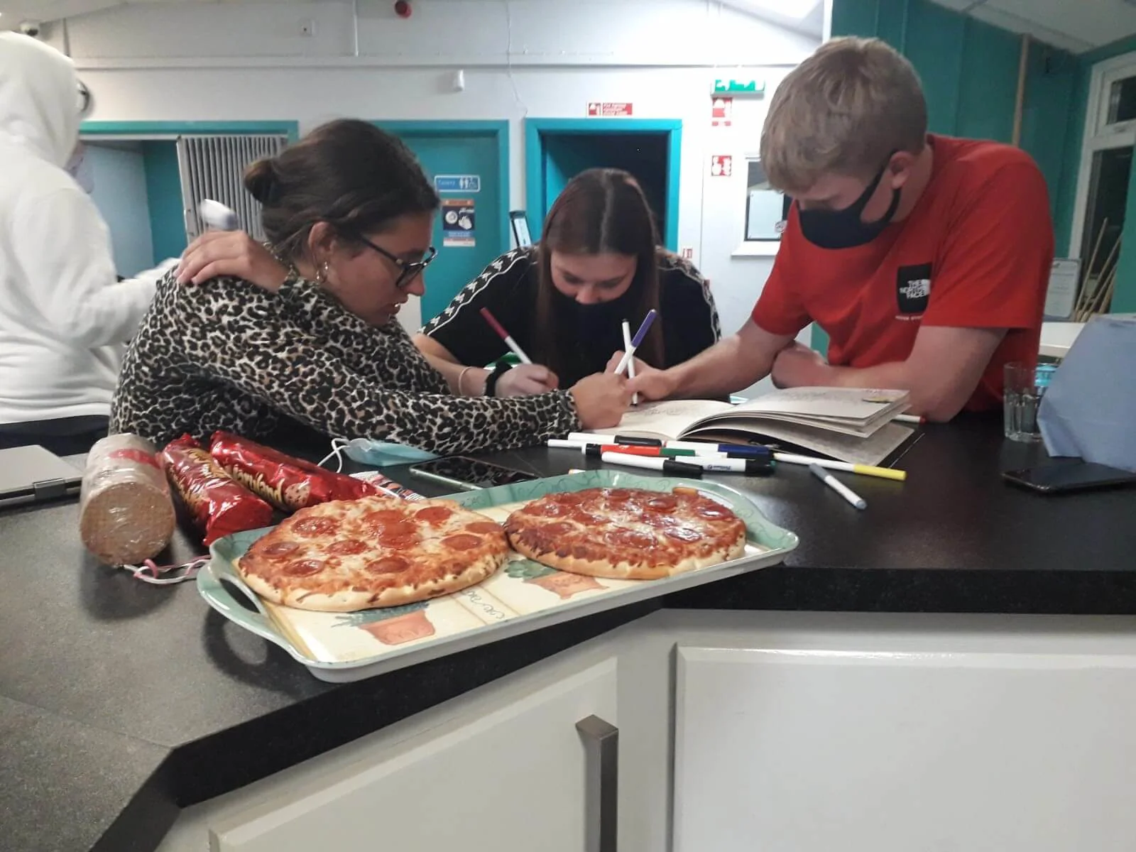 A youth club leader and two young people drawing with felt tips, with pizza on the counter beside them