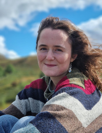 Photo of Maggie sitting outside. She has brown hair, is smiling and wearing a stripey jumper