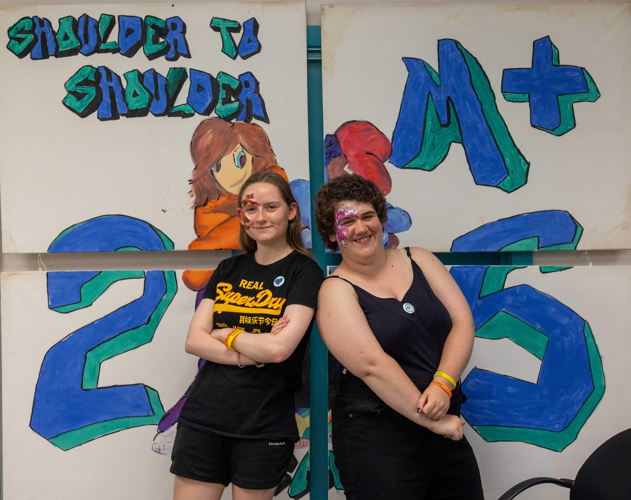 Two young people posing in front of a Shoulder to Shoulder 25 anniversary backdrop