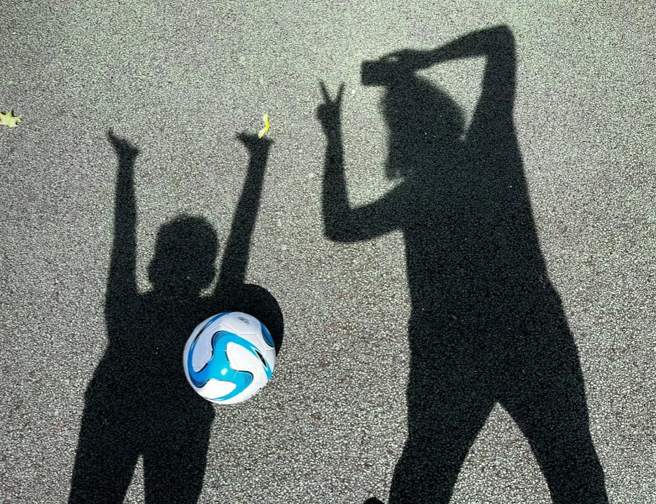 Shadows of two people with arms raised playfully on tarmac, with a football on the ground between them