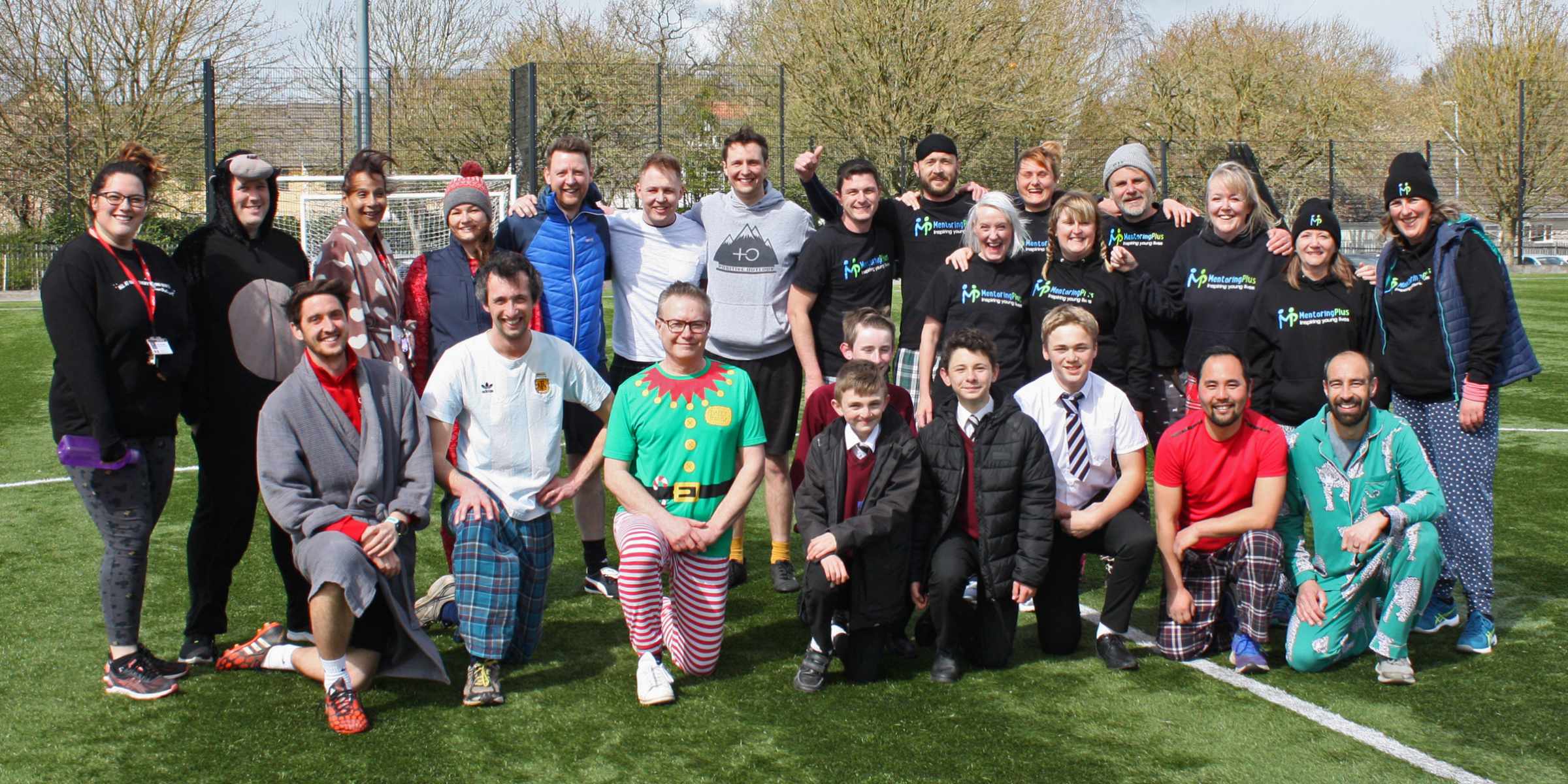 A large group in fancy dress and pyjamas posing together on a football pitch at a Mentoring Plus fundraising football event